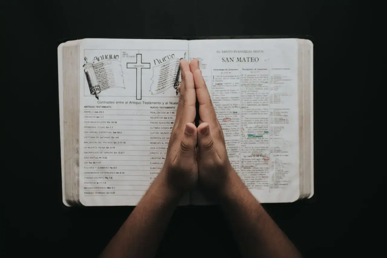 Dłonie złożone do modlitwy nad otwartą Biblią. Pamiętaj człowiecze na Jezusa, szukaj Jego światła w Słowie.
