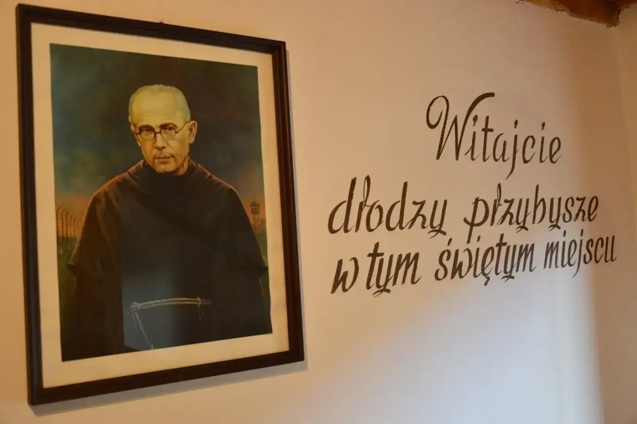 Obraz przedstawia portret błogosławionego Maksymiliana Kolbe w habicie, z widokiem na ob&oacute;z koncentracyjny w tle. Obok napis: 